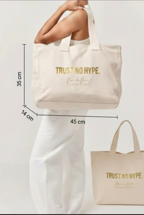 Model holding a large beige canvas tote bag with gold "TRUST NO HYPE" text, showing the product's size and fit.