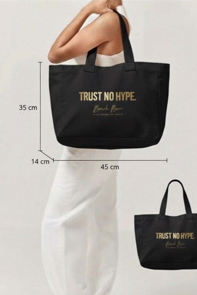 Model holding a large black canvas tote bag with gold "TRUST NO HYPE" text, showing the product's size and fit.