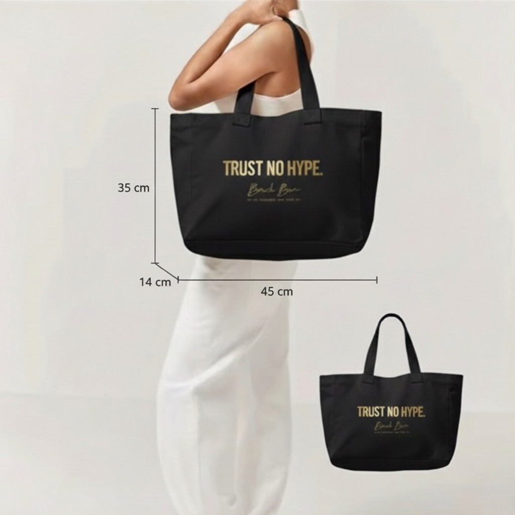Model holding a large black canvas tote bag with gold "TRUST NO HYPE" text, showing the product's size and fit.