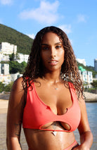 A woman wearing a red bikini top with cut-out details and removable padding, standing by the water.