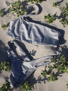 A silver one shoulder bikini top with an asymmetrical shoulder strap, laid out on a sandy surface with foliage around.