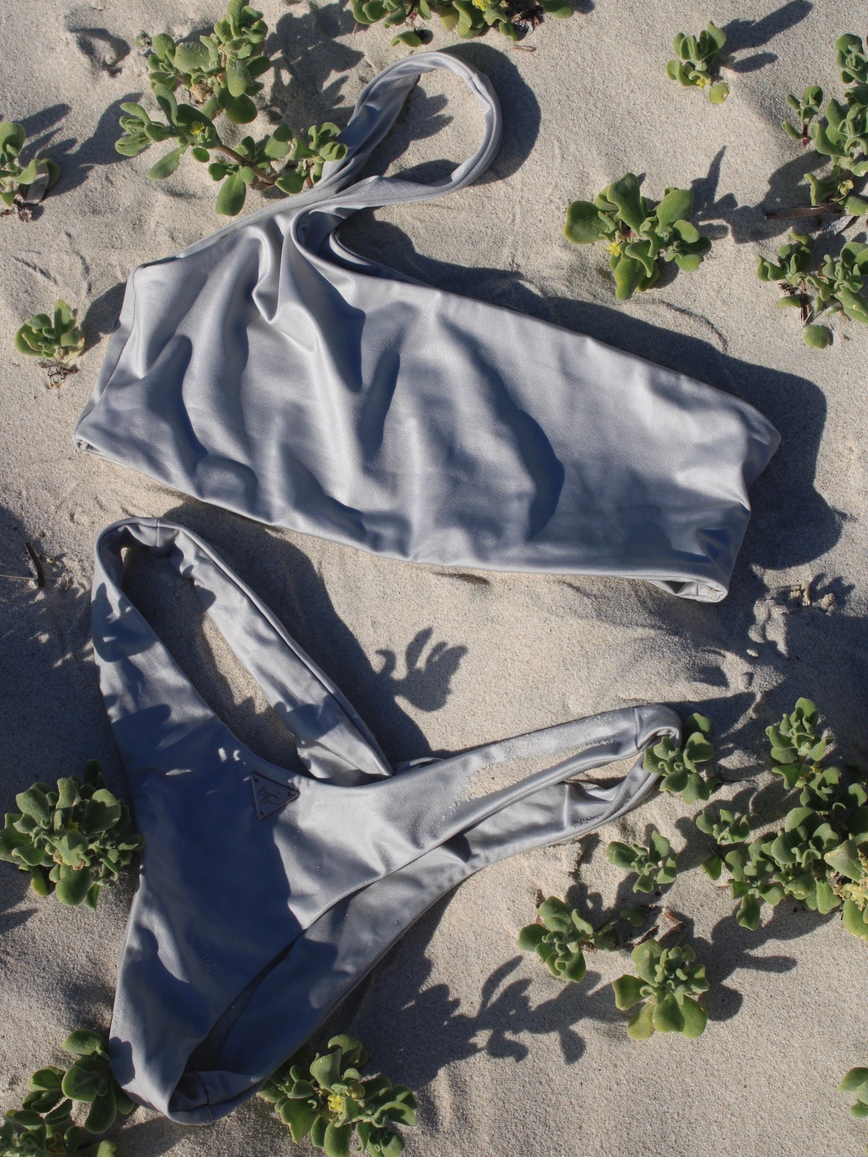 A silver one shoulder bikini top with an asymmetrical shoulder strap, laid out on a sandy surface with foliage around.