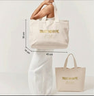 Model holding a large beige canvas tote bag with gold "TRUST NO HYPE" text, showing the product's size and fit.