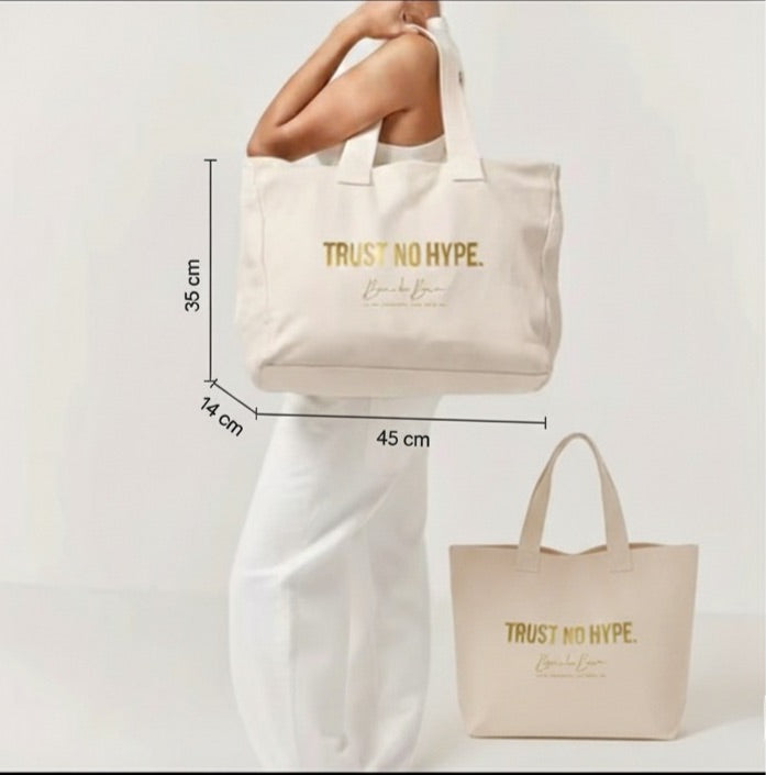 Model holding a large beige canvas tote bag with gold "TRUST NO HYPE" text, showing the product's size and fit.