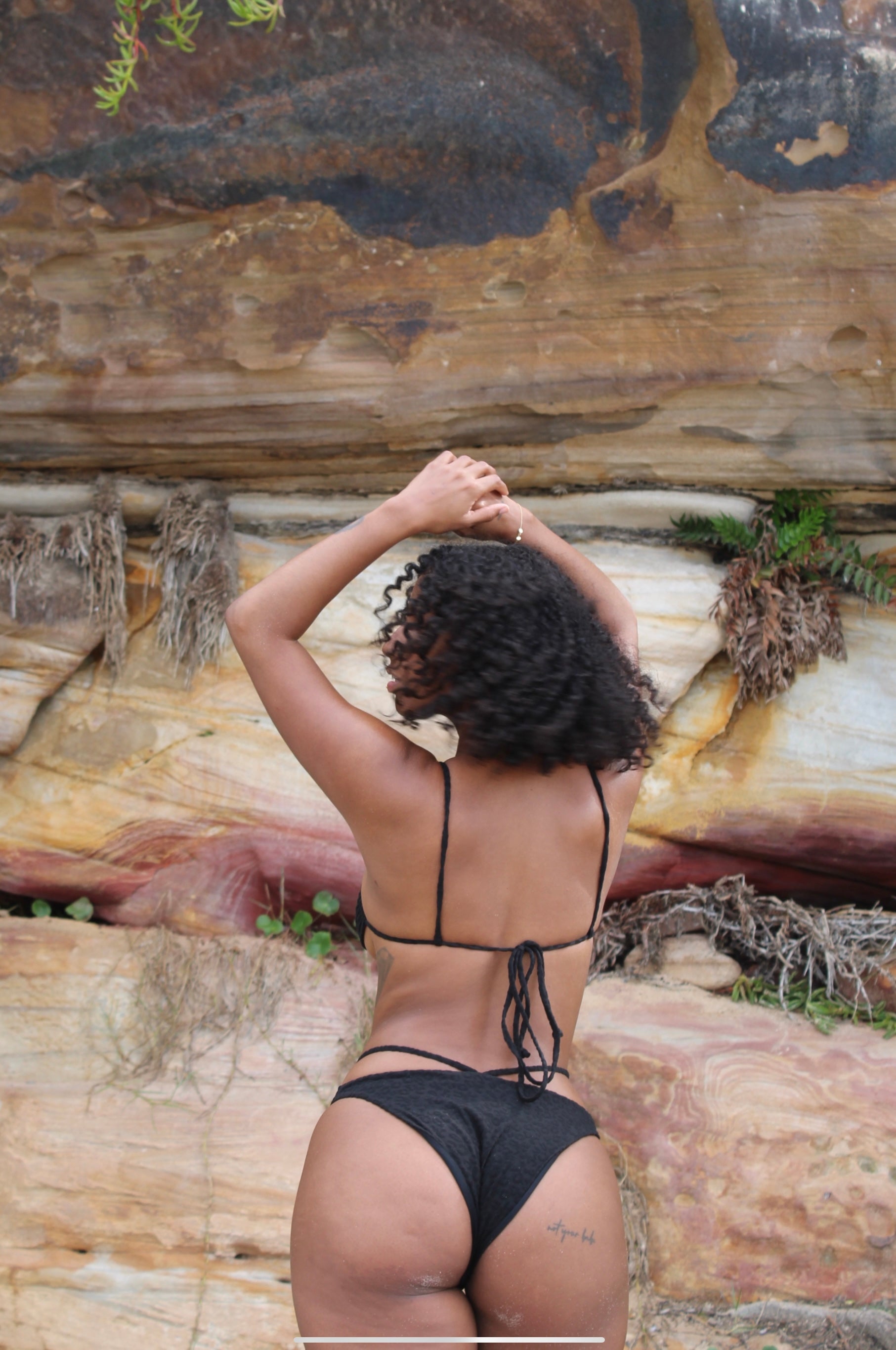 A woman wearing a black textured bikini being showed from the back infront of a big stone.