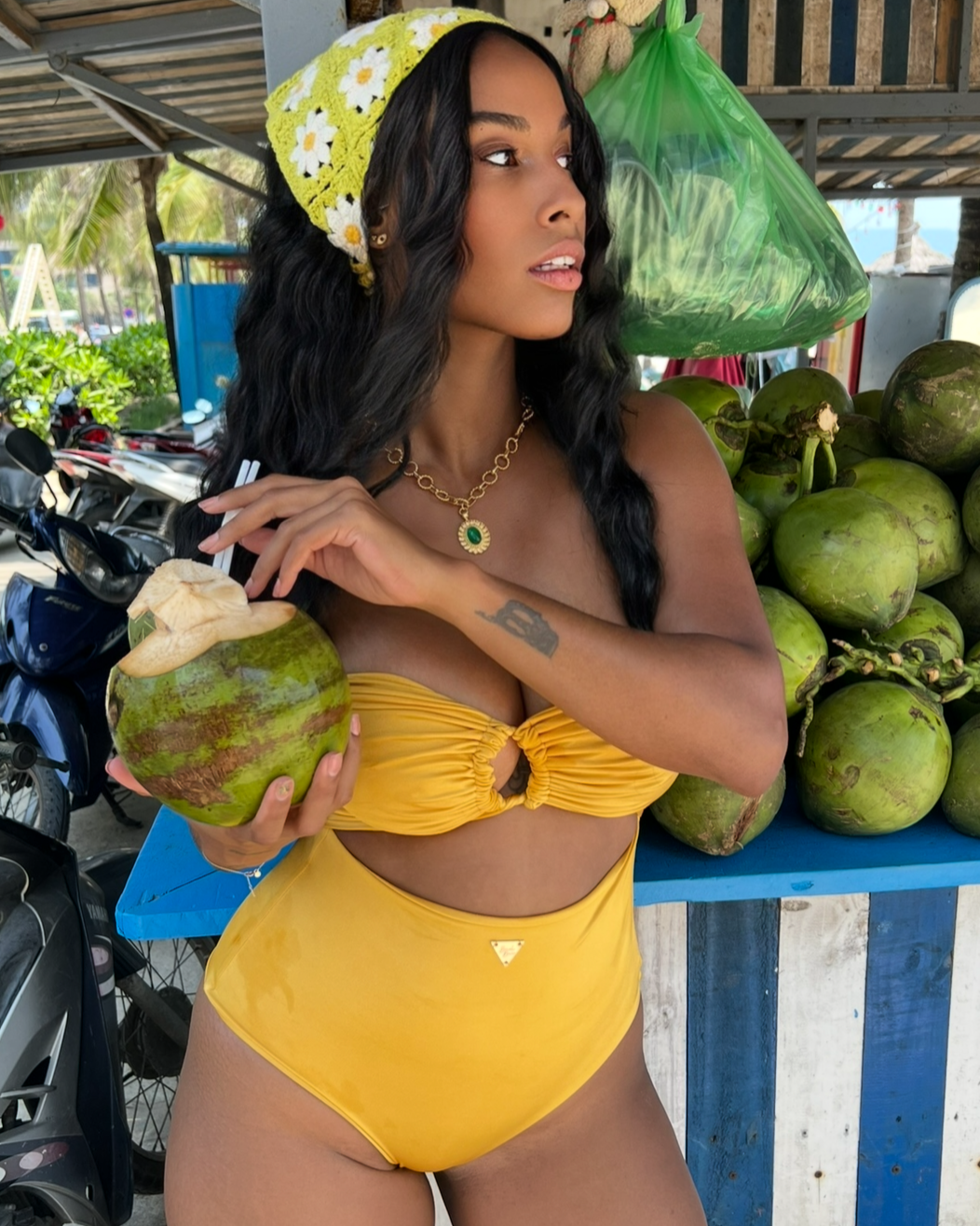 Woman in a yellow swimsuit holding a green coconut in an outdoor setting with motorcycles and a table of coconuts.