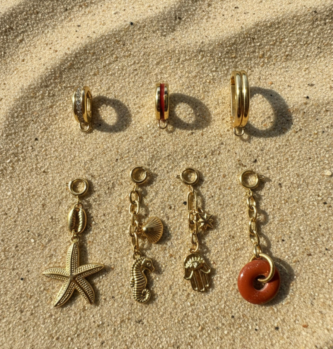 Set of gold charm bikini clips with various charms on a sandy surface