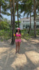 A girl in a pink bikini holding a mango shake walking away from pal trees and to the beach.
